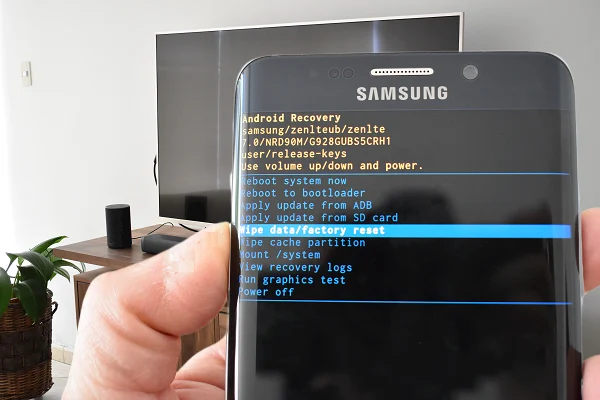 La imagen muestra un teléfono Samsung con la pantalla para hacer un reseteo de fábrica. En el fondo se aprecia un smart tv sony y un altavoz Amazon Echo.